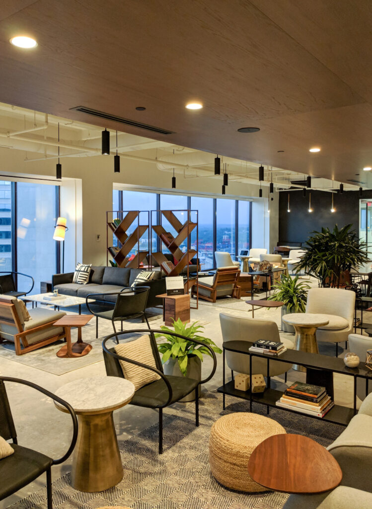 Modern lounge with varied seating, small side tables, a large wooden bookshelf, and floor-to-ceiling windows overlooking a city skyline.
