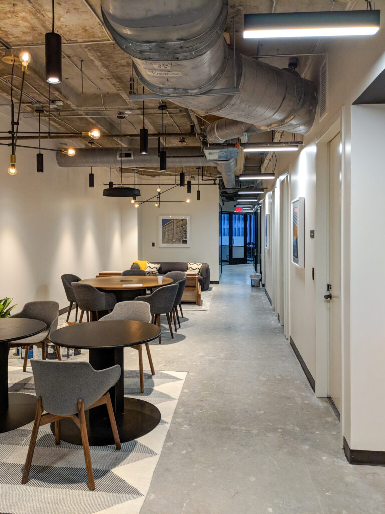 An open office area with small round tables, grey chairs, and a seating area with a sofa, featuring exposed industrial ceiling ducts and pendant lighting.