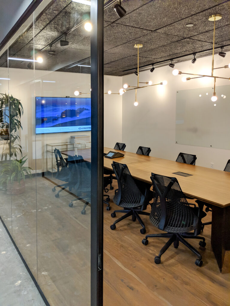 A glass-walled conference room featuring a long wooden table, black office chairs, a mounted display screen, and a wall-mounted whiteboard.