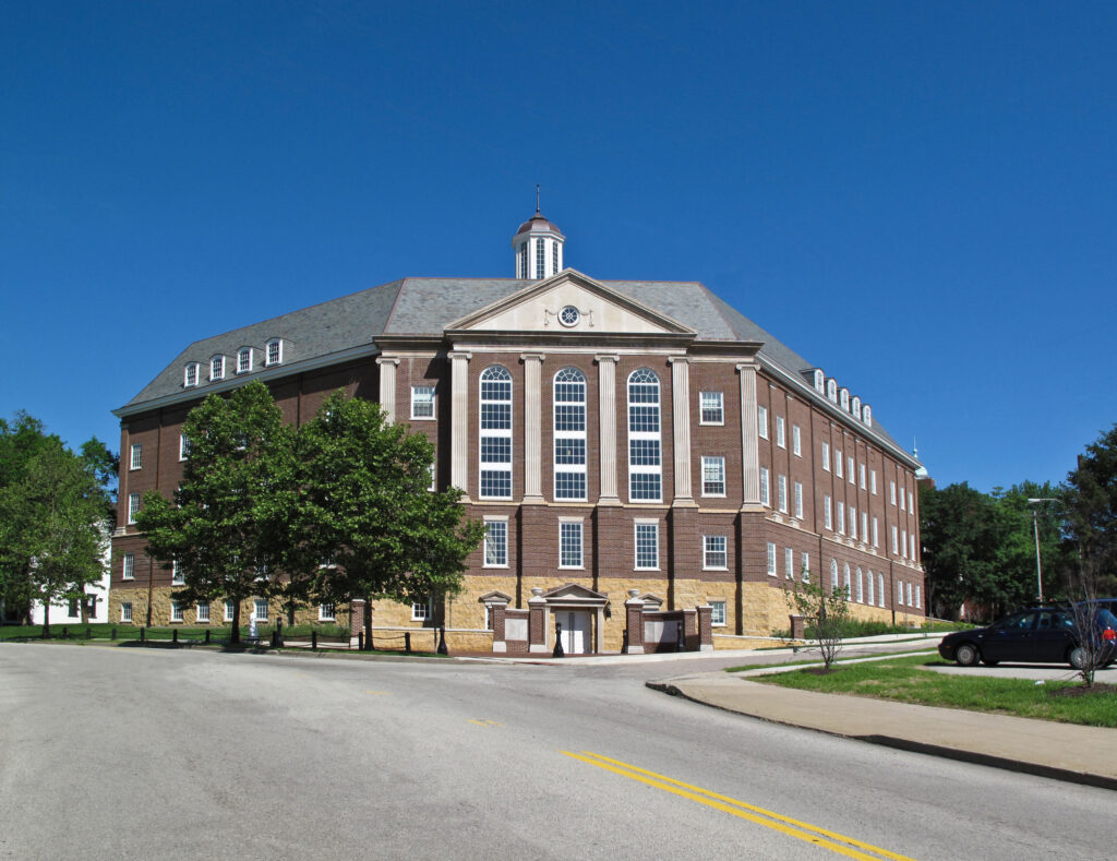 A large, multi-story brick academic building with white columns and a central cupola under a clear blue sky.
