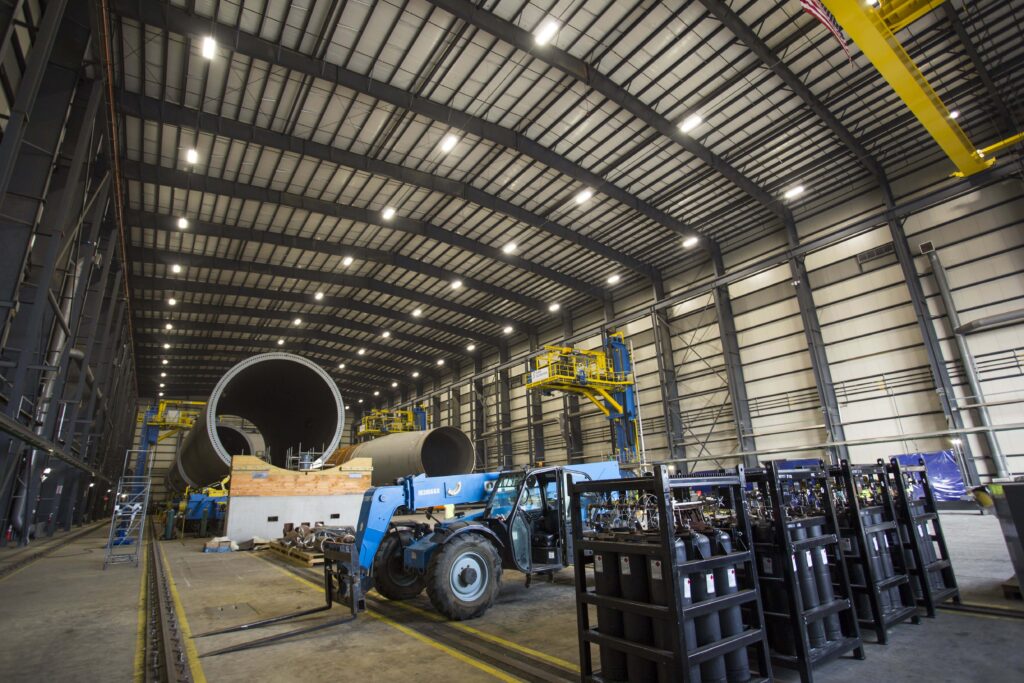 Large cylindrical steel sections inside a high-ceilinged industrial manufacturing facility, with a telehandler and racks of gas cylinders in the foreground.