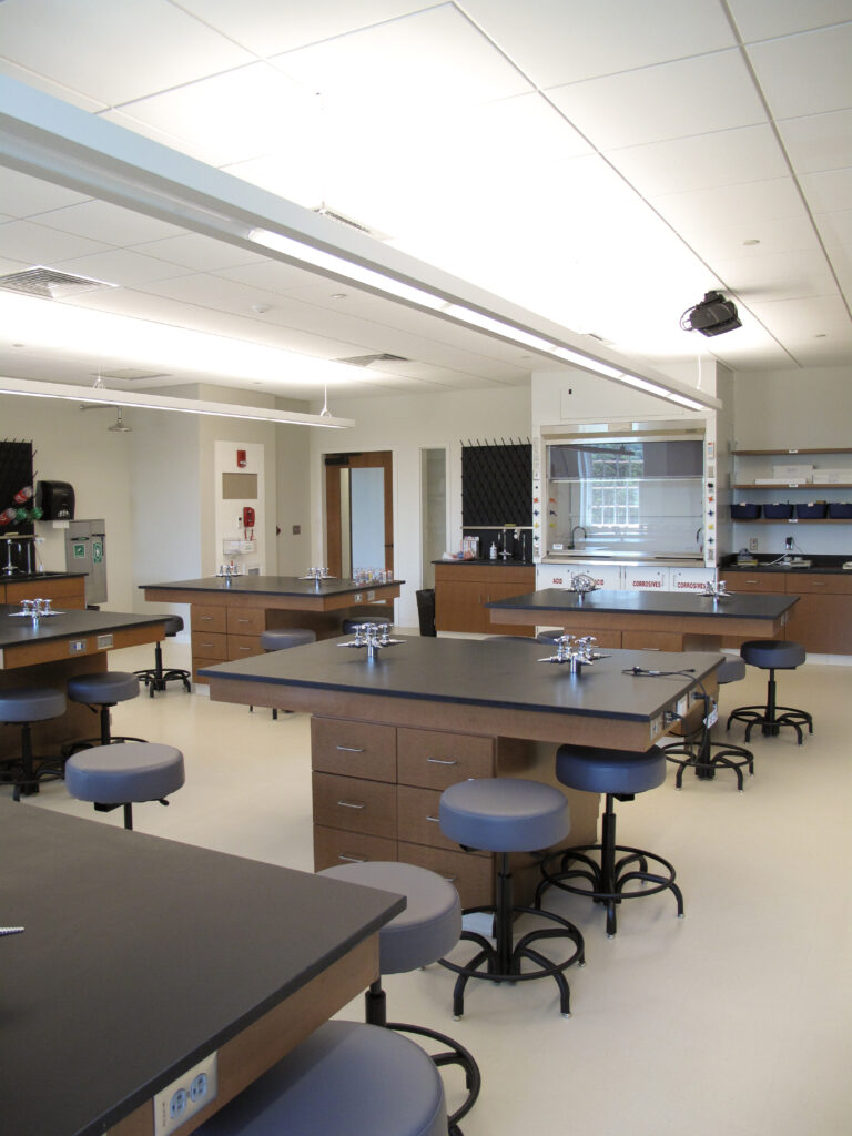 A science laboratory classroom with black-topped work tables, stools, and a fume hood.