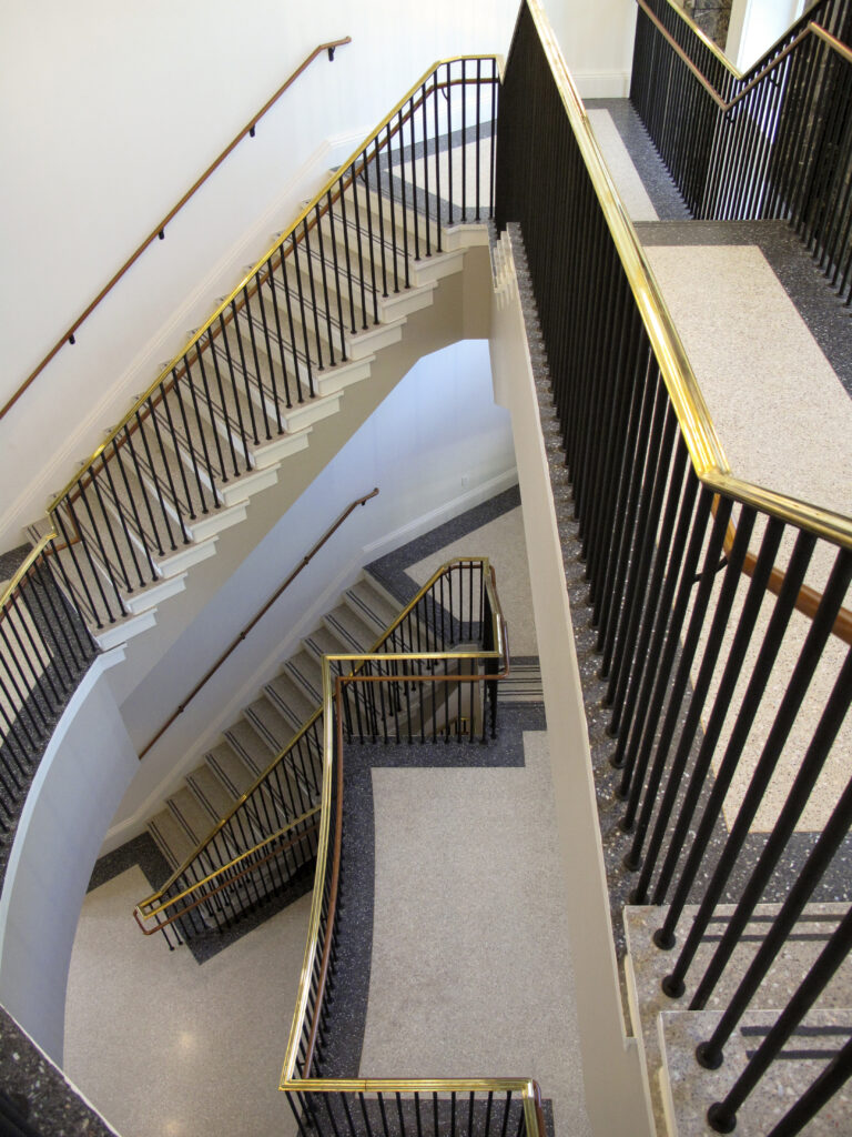 A multi-level staircase with light-colored stone steps, dark metal railings, and brass handrails viewed from above.