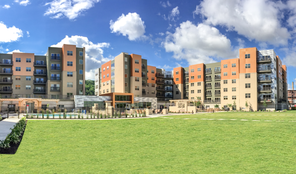 A large, multi-story apartment complex with a tan, orange, and gray facade overlooking a grassy lawn and an outdoor swimming pool.