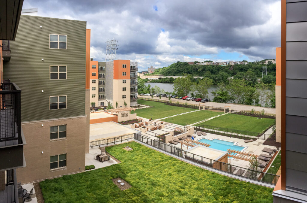 High-angle view of an apartment complex courtyard with a swimming pool, lounge seating, and a green roof, overlooking a river and hillside.