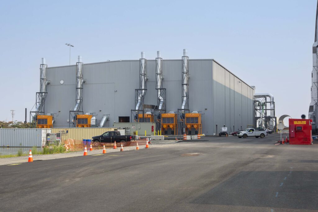 A large industrial metal building with multiple exterior exhaust stacks and orange machinery units, set in a paved lot with a few parked pickup trucks.