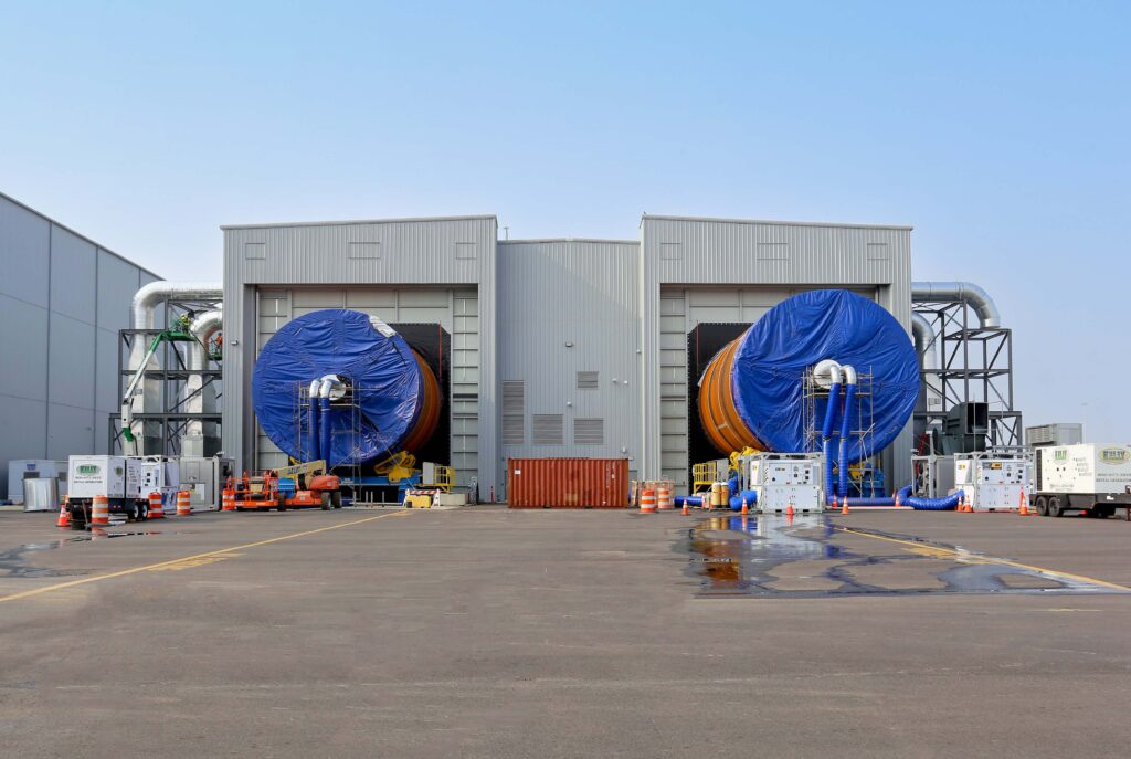 Two large industrial machines covered in blue tarps positioned outside an aerospace facility.