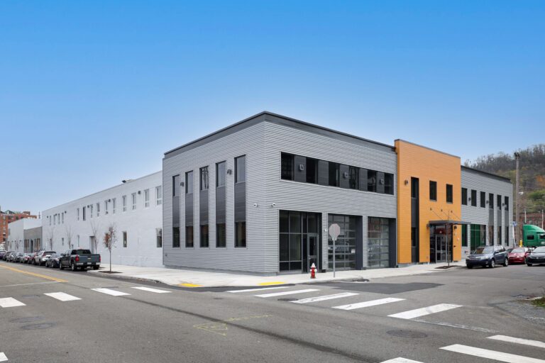 A street-level view of a multi-story commercial building with grey corrugated metal and orange accent panels, situated at a corner intersection with parked cars.