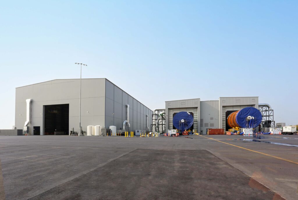 Two large, cylindrical rocket components covered in blue tarps sit outside the bays of a hangar building.