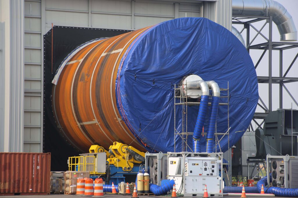 Large cylindrical orange rocket stage partially covered by a blue tarp, sitting on a yellow transport platform in front of a facility.