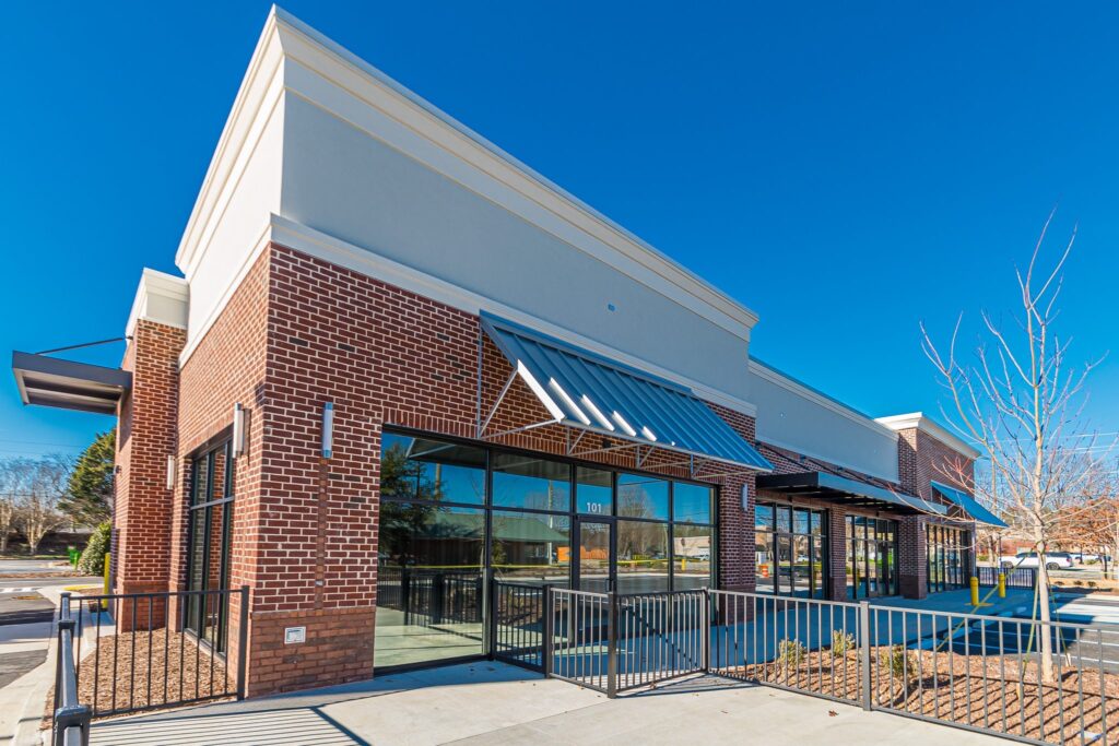 A modern, one-story brick retail building with large glass storefronts, metal awnings, and a black metal railing along the front walkway.