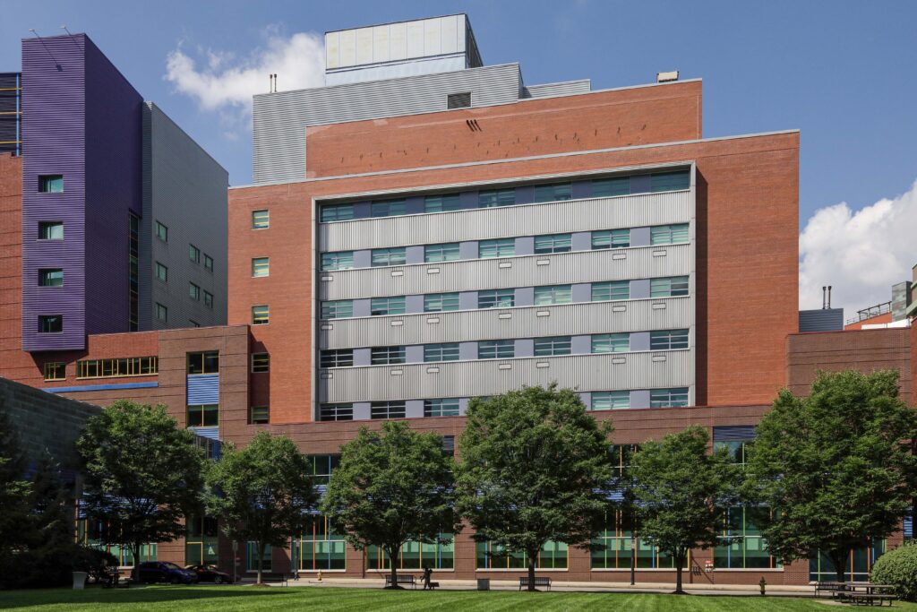 Large academic building with brick and metal panel facades, viewed from a grassy lawn with trees in front.
