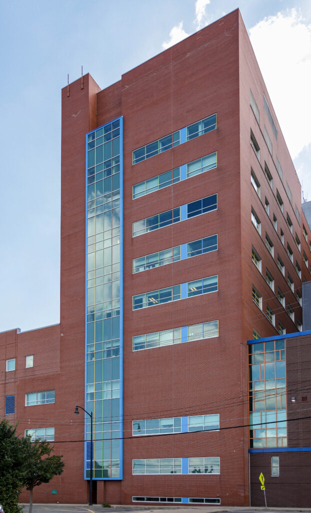 A tall red brick building with a vertical blue glass window section and a pedestrian crossing sign on the sidewalk.