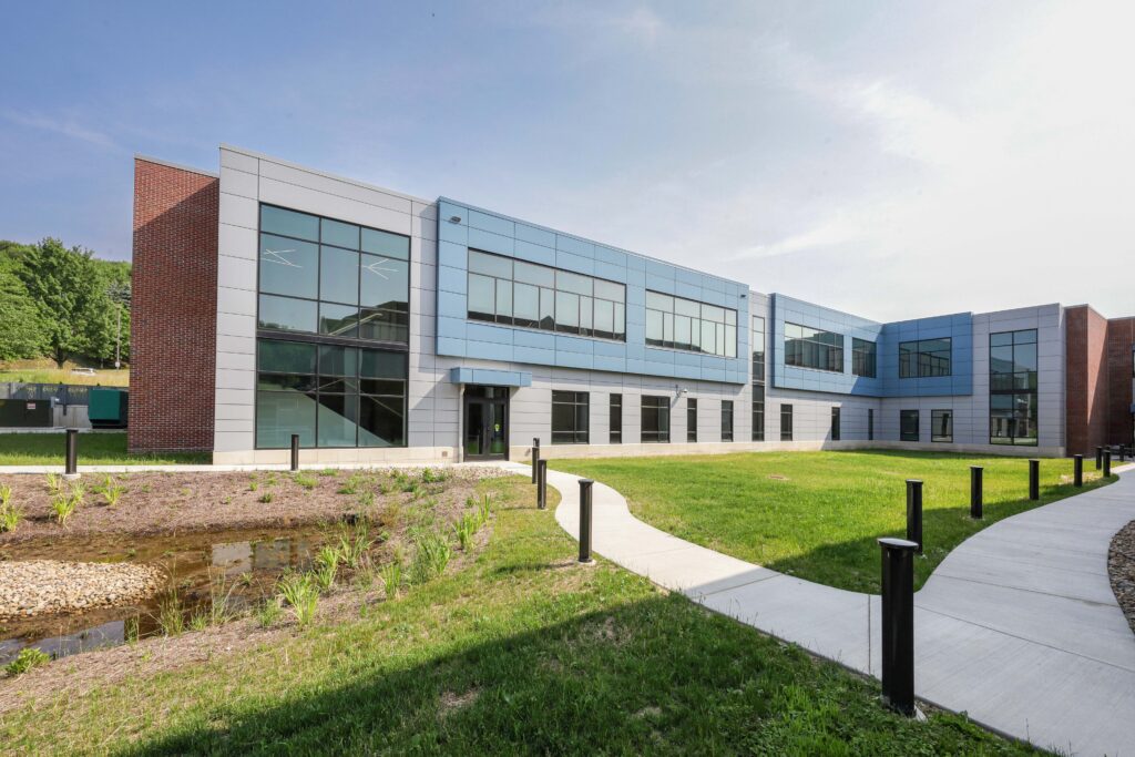 A modern two-story building with blue and gray panels and large windows, fronted by a grassy lawn, a small pond, and a curved concrete walkway.