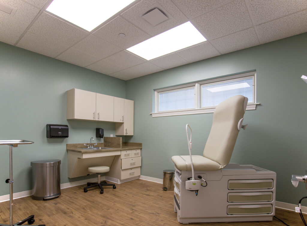A medical examination room with an adjustable exam table, a sink, wall-mounted cabinets, and a rolling stool.