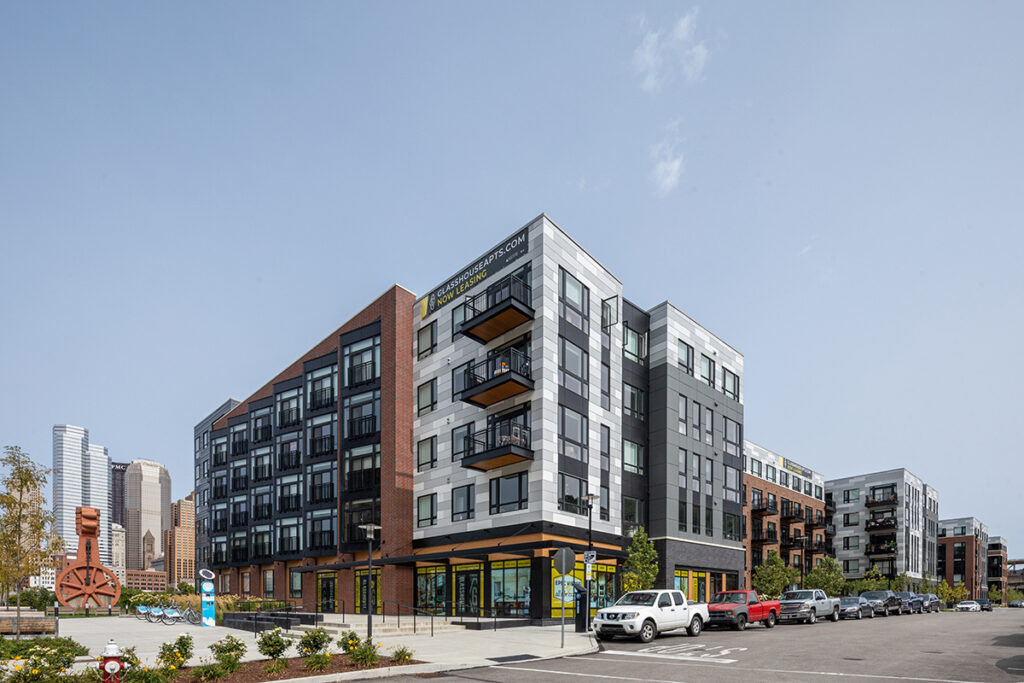Multi-story apartment building with a “Now Leasing” sign, ground-floor commercial space, and parked cars, with a city skyline in the distance.
