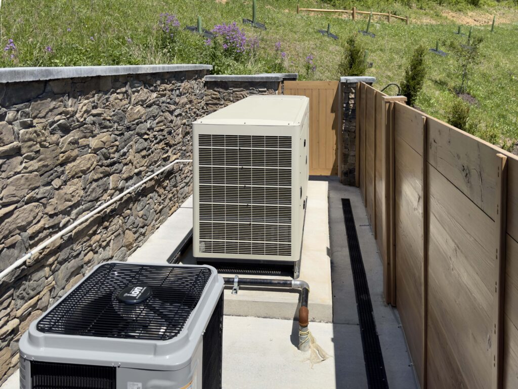 Large beige backup generator on a concrete pad, next to a smaller grey air conditioning unit, situated between a stone retaining wall and a wooden privacy fence.