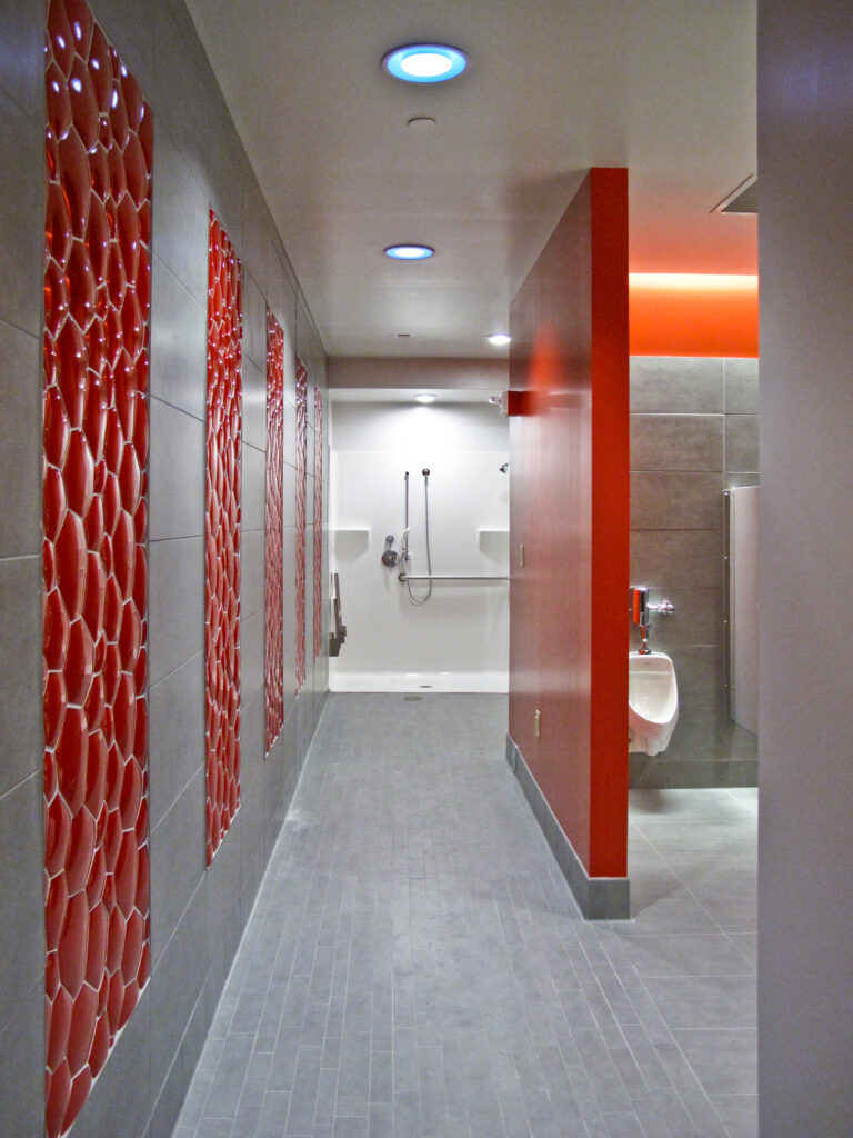 Modern public restroom with a row of red decorative wall panels, a urinal on a red partition wall, and an accessible shower stall at the end of the corridor.