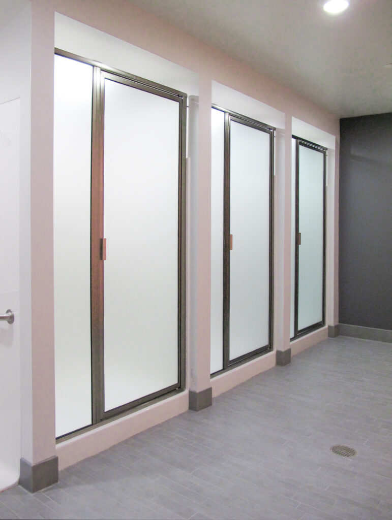 Three shower stalls with frosted glass doors and dark metal frames lined up along a tiled wall.