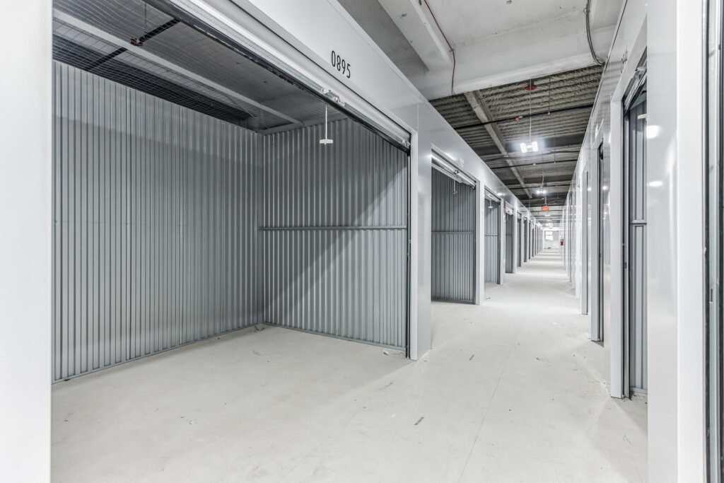 A hallway in a storage facility lined with empty storage units featuring corrugated metal roll-up doors.