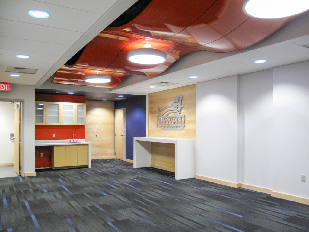 Interior view of a modern room with a dark carpet, a small kitchenette, and a wall featuring the Duquesne University basketball logo.