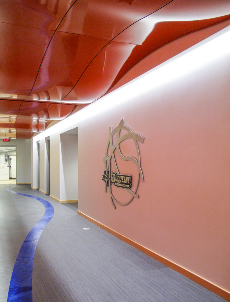 A hallway featuring a Duquesne Basketball metal wall logo and a curved blue floor stripe under a glossy red ceiling.