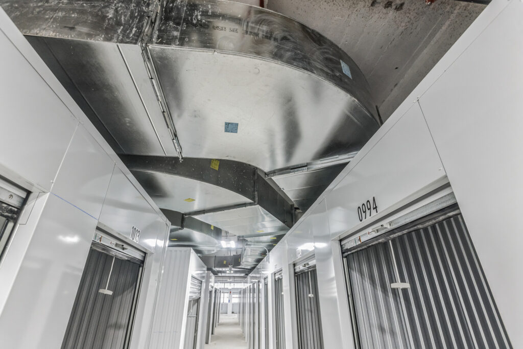 A hallway of storage units with metal roll-up doors, labeled with unit numbers, beneath exposed ceiling ductwork.