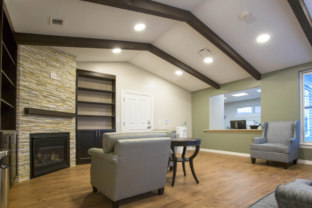 A waiting area with a stone fireplace, built-in shelving, two armchairs, and a round side table, viewed through an interior window.