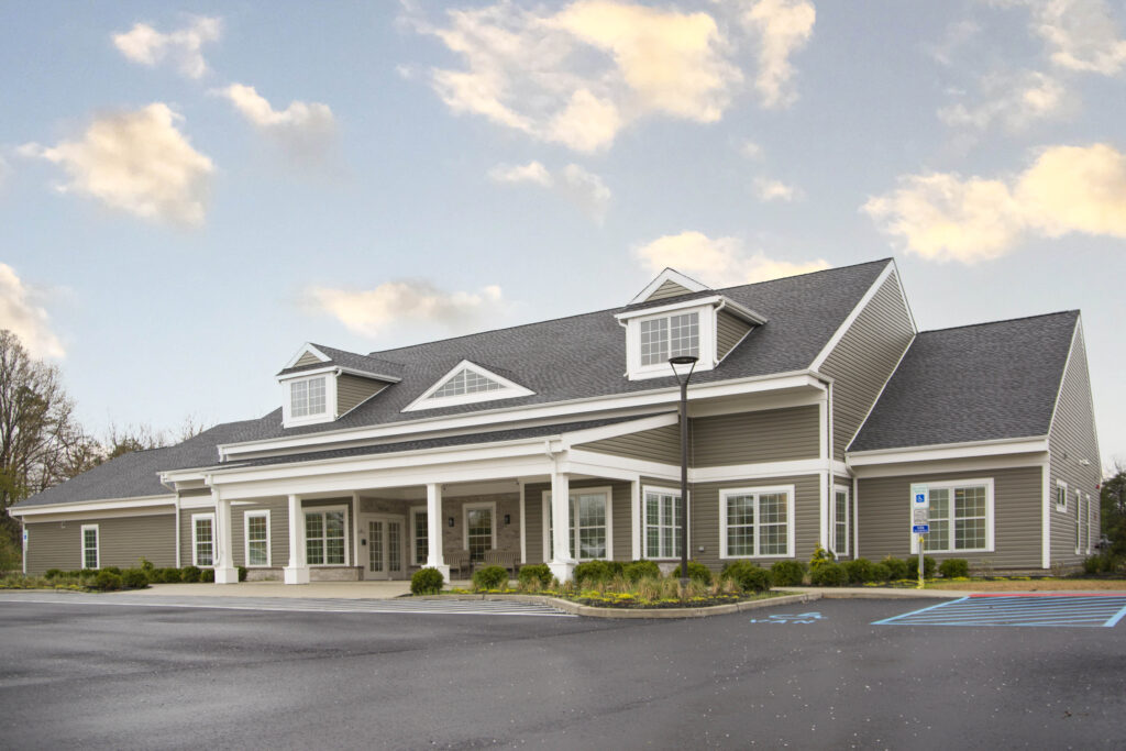 A single-story commercial building with grey siding, a dark shingled roof, and a front portico, seen from a paved parking lot.