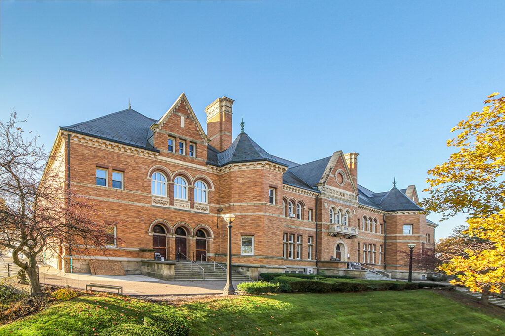 A historic three-story brick building with arched windows and a slate roof, set against a clear blue sky.