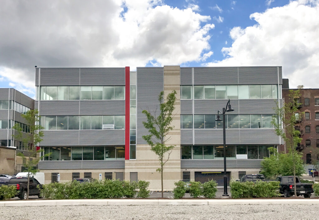 A three-story modern office building with gray metal siding, a vertical red accent, and a light-colored stone column, with parked cars in the foreground.