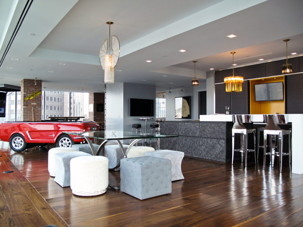 A modern lounge featuring a red vintage car base as a table, a glass dining table with tufted ottomans, and a bar area with stools.