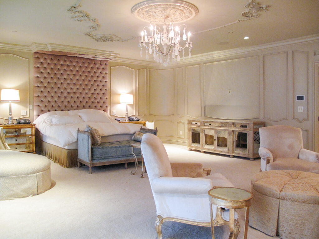 A bedroom with a tufted pink headboard, a chandelier, and pale neutral furniture, including a bed, an upholstered bench, two armchairs, and matching ottomans.