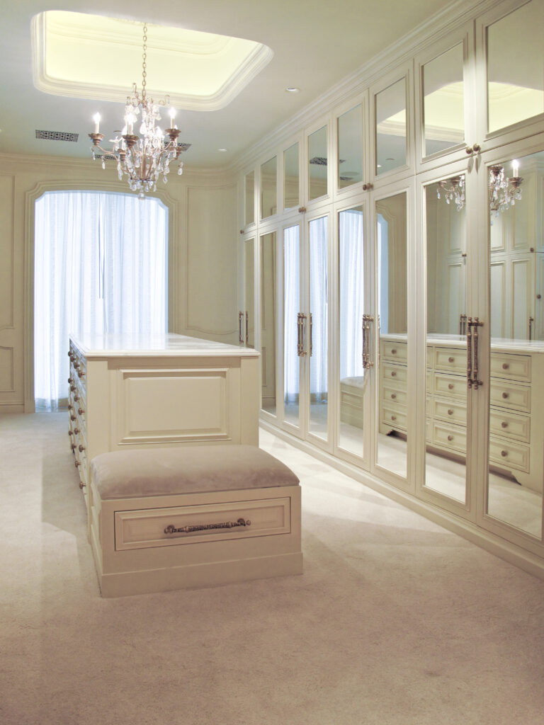 A walk-in closet featuring a central island with a bench, surrounded by floor-to-ceiling mirrored cabinetry and a crystal chandelier.