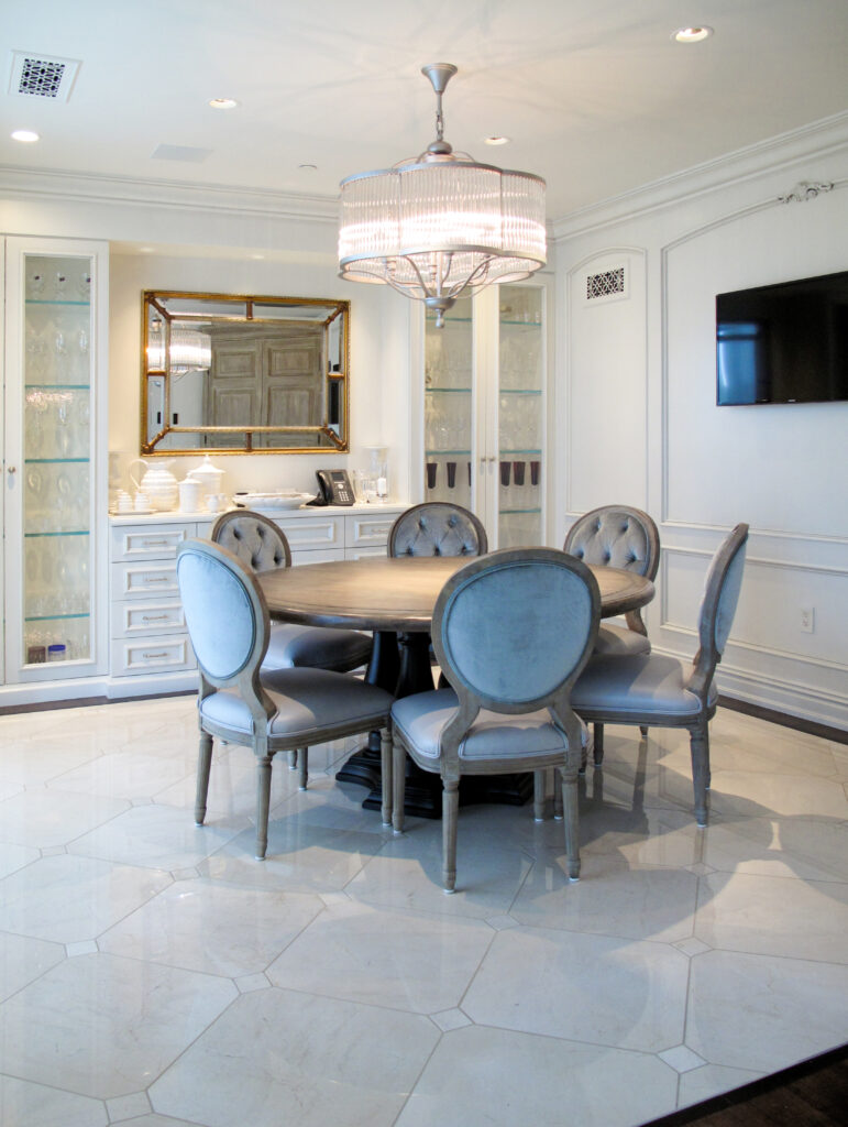 A round wooden dining table surrounded by six tufted light blue upholstered chairs in a room with a crystal chandelier and glass-front cabinets.