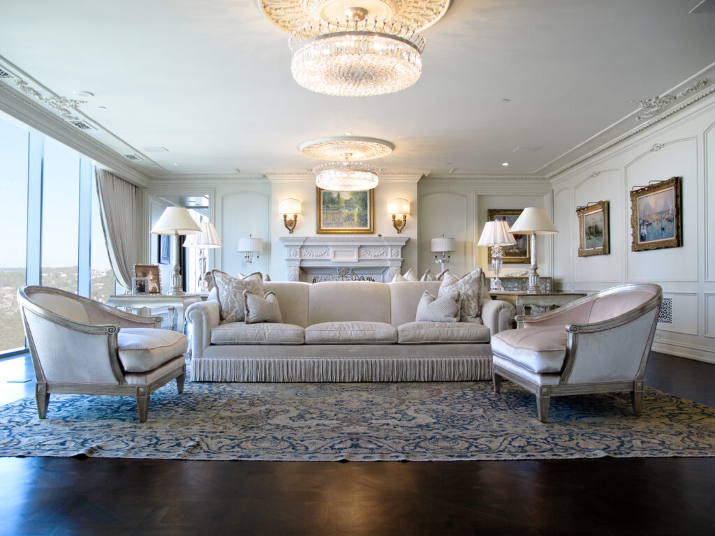 A living room featuring a long, cream-colored sofa flanked by two matching armchairs, arranged on a patterned rug in front of a fireplace with two crystal chandeliers above.