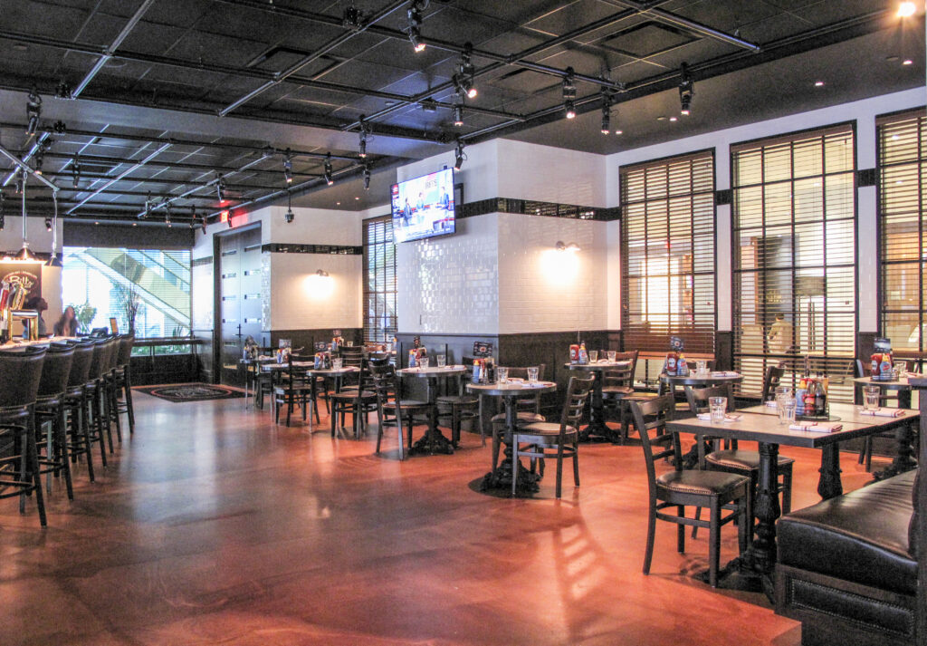 Interior of a restaurant with a bar counter on the left, several dining tables and chairs arranged throughout the room, and a television mounted on a tiled wall.