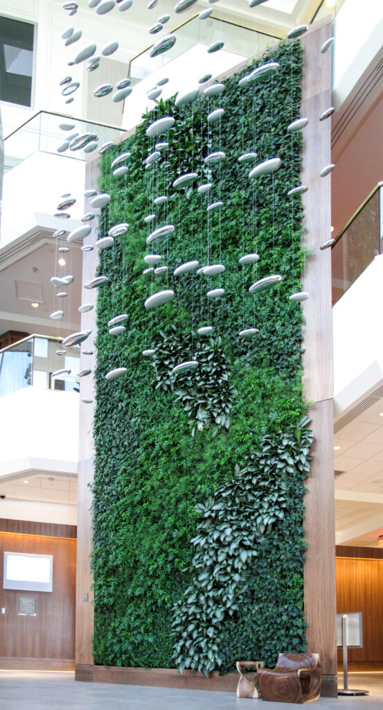 A tall indoor living plant wall decorated with hanging metallic, pebble-shaped sculptures in a multi-story atrium.