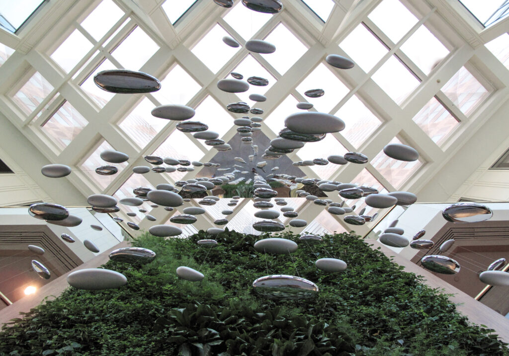 A ceiling installation of many mirrored, oval-shaped sculptures suspended over a large indoor living wall.