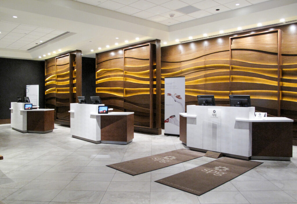 Hotel reception area with three white and wood-paneled service counters in front of a decorative, illuminated wave-patterned wall.