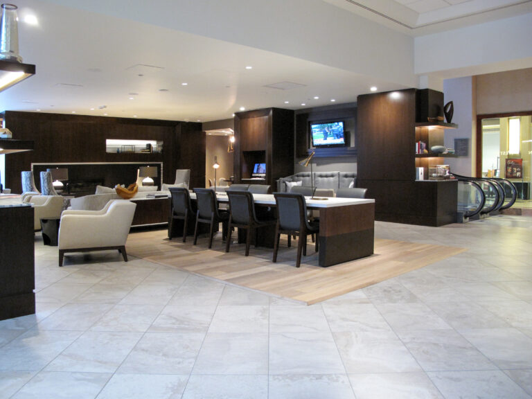 A hotel lobby lounge featuring a long dining table, seating areas with armchairs and sofas, and an escalator in the background.