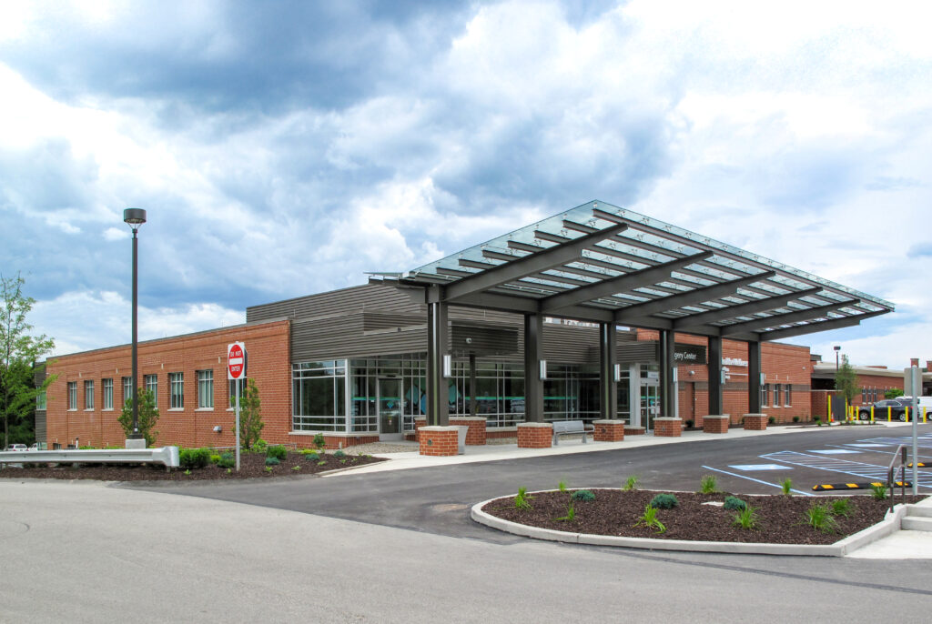 A low-rise brick medical building with a large glass canopy over the entrance and a parking lot in the foreground.