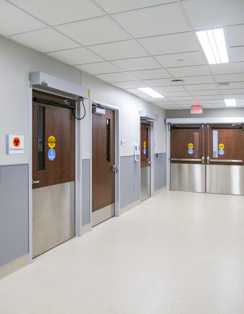A hospital hallway with several wooden doors, including a Decontamination room door and a set of double exit doors.
