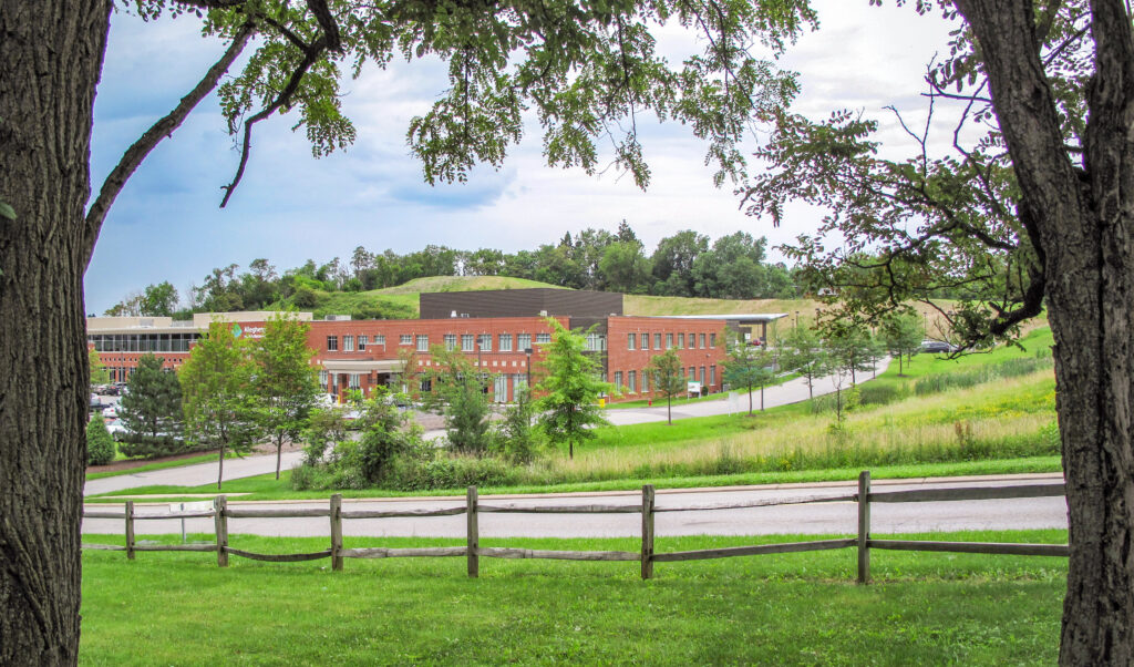 A red brick building with a parking lot, viewed across a grassy field with a wooden split-rail fence in the foreground.