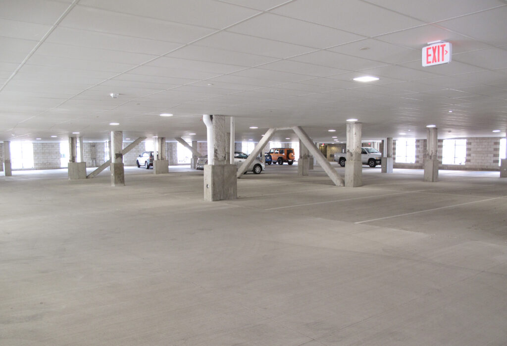 A mostly empty parking garage with concrete support columns, a few parked cars, and an illuminated red exit sign.