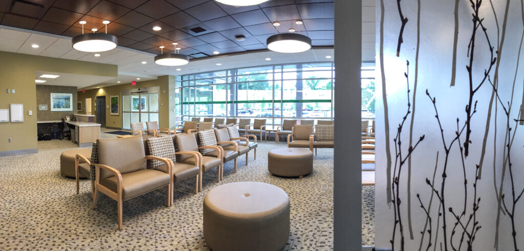 A panoramic view of a medical waiting room with rows of chairs, circular ottomans, and large windows.
