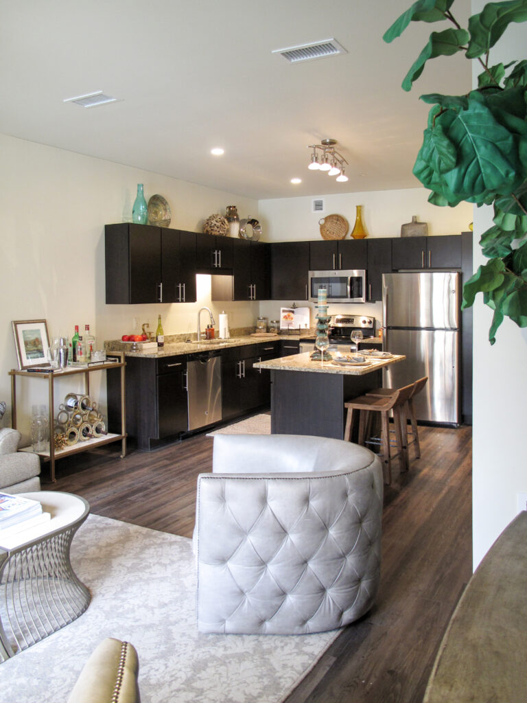 A kitchen with dark cabinets, stainless steel appliances, and a central island, viewed from a living area featuring a tufted armchair and a bar cart.