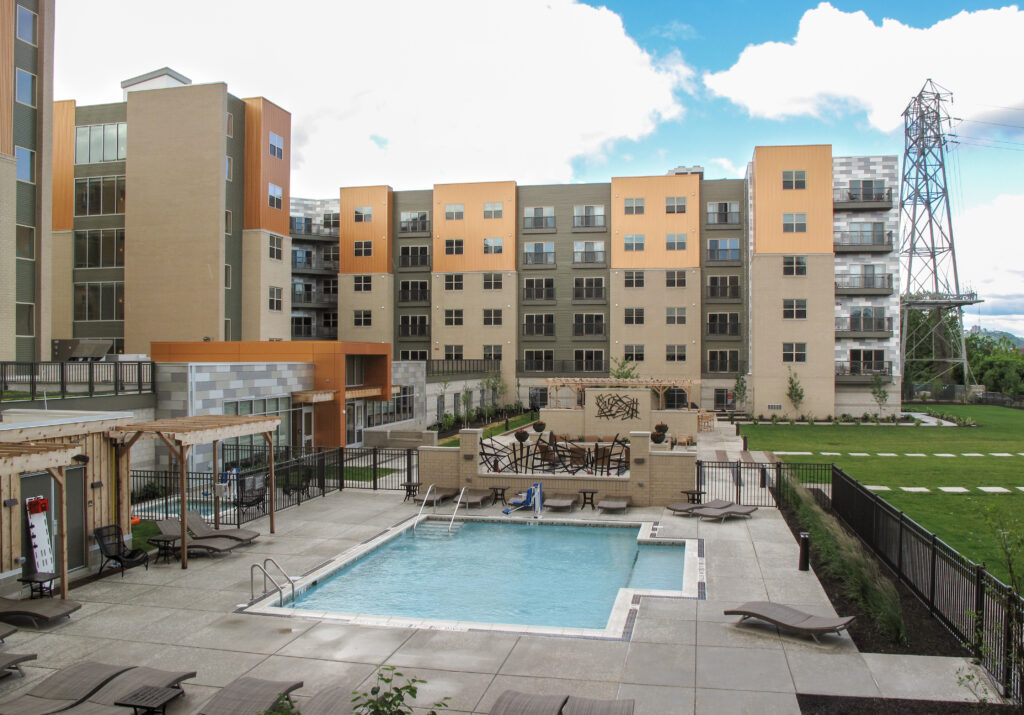 A multi-story apartment complex with a central courtyard featuring a swimming pool, lounge chairs, and a wooden pergola.