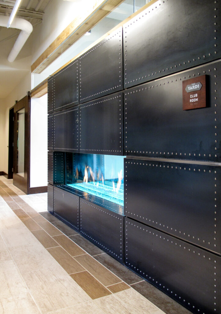 A linear fireplace built into a black, rivet-paneled wall, with a sign reading “The Yards Club Room.”