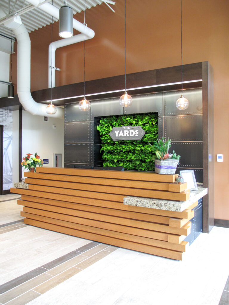 Reception desk with a horizontal wood-slat front, positioned in front of a green plant wall featuring a sign that reads “The Yards.”